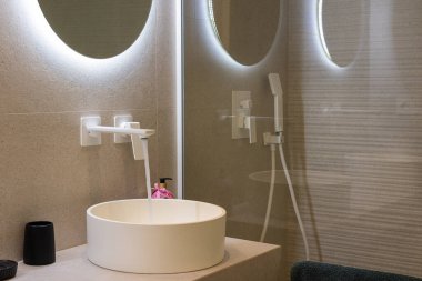 Yuvarlak ışıklı aynalı modern banyo, beyaz musluk, ve taş döşeli duvarlara karşı parlak yuvarlak bir havuza akan su..