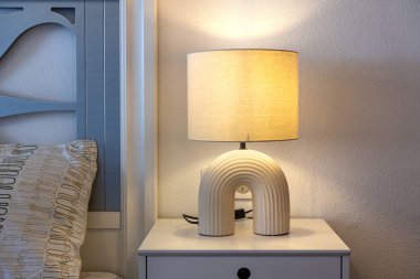 Modern bir komodinin üzerinde kumaş gölgesi olan şık bir krem masa lambasının yakın plan resmi..