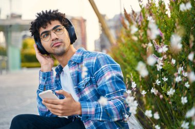 Dalgalı kıvırcık saçlı hippi adam elinde cep telefonu, kablosuz kulaklıkla müzik dinliyor başka tarafa bakıyor, parkta oturuyor. Modern teknoloji kavramı  