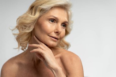 Kendine güvenen, kendine güvenen, saçları dalgalı, yüzüne dokunan, arka planda izole bir şekilde duran olgun bir kadın. Cilt bakımı kavramı