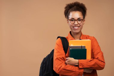 Gözlüklü gülümseyen Afrikalı Amerikalı üniversite öğrencisi bej arka planda izole edilmiş kitap ve sırt çantası tutuyor. Eğitim kavramı 