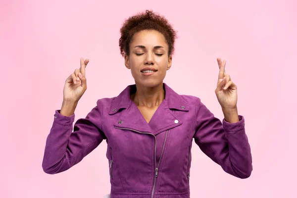 Beautiful stylish African American woman with fingers crossed and eyes closed wish something, hopes for luck  isolated on pink background