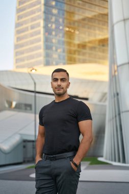 Kenar mahallede duran, siyah günlük kıyafetler giyen, kendine güvenen orta doğulu adamın portresi.                           