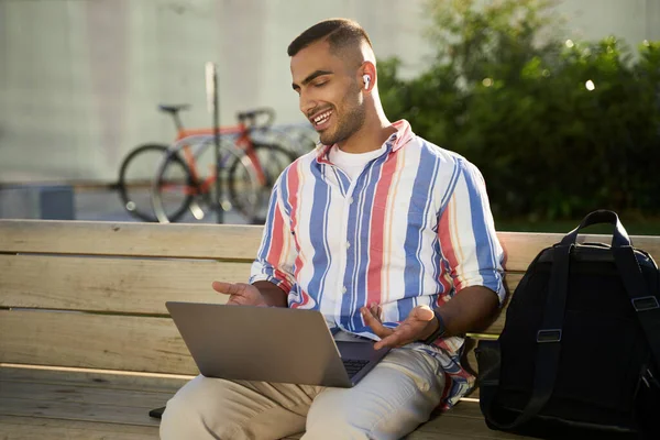 Handsome smiling middle eastern man using laptop computer having video call sitting on bench. Young successful freelancer working online at workplace. Remote job concept