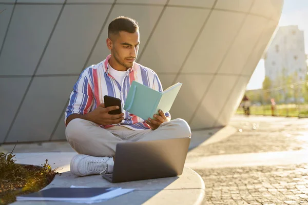 Pensive middle eastern student studying, exam preparation, reading book in university campus. Modern Iranian man using laptop, holding mobile phone working sitting at workplace