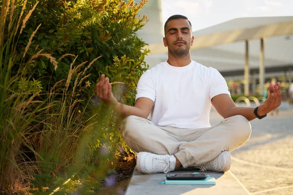 Young handsome Iranian man with closed eyes meditation sitting in lotus pose, relaxing in park. Yoga, healthy lifestyle concept