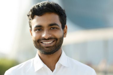 Üniversite kampüsünde kameraya bakan beyaz gömlekli, gülümseyen Hintli bir öğrencinin yakın plan portresi. Eğitim kavramı