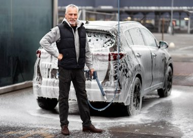 Handsome elderly man, expressing positive emotions, smiling toothy smile, looking at camera, posing with high pressure hose against car full of cleaning foam, in self-service manual touchless car wash