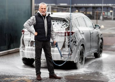 Handsome elderly man, expressing positive emotions, smiling toothy smile, looking at camera, posing with high pressure hose against car full of cleaning foam, in self-service manual touchless car wash clipart