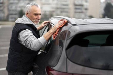 Waist-up side portrait of a Caucasian gray-haired senior man, cleaning car with microfiber cloth, at car wash self service outdoors. The male driver applying wax and polishing scratches with a towel
