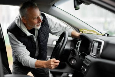 Handsome Caucasian senior active male driver, cleaning his car at self service car wash. He's wiping interior, steering wheel and dashboard with yellow microfiber cloth, waxing and polishing scratches