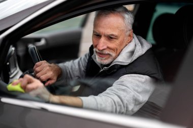 Close-up portrait of positive Caucasian 60-70 years old man cleaning car interior with yellow microfiber cloth. The concept of car maintenance, detailing, valeting and car wash at self-service station