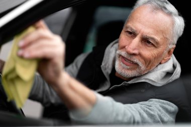 Close-up portrait of handsome positive white-bearded Caucasian 60-70 years old man, sitting on the driver's seat and wiping the car's side mirror. Car wash and auto service concept