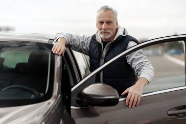 Close-up portrait of a Caucasian confident senior man in casual wear, standing by his new car in the outdoors parking lot of a dealership. Buying new automobile. Driving Lifestyle and People concept