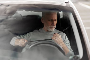 Caucasian gray-haired senior man driver, sitting on driver's seat and fastening the seat belt in the car before a trip by ride. Driving safety and compliance with traffic rules