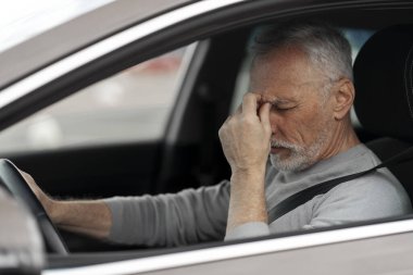 Overworked elderly Caucasian man 60-70 years old, taxi driver feeling headache and tired, stopping after driving a car in a traffic jam. Close-up of an exhausted senior male driver