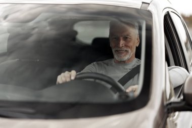 View through the windshield to a Caucasian smiling 60-70 years senior man with fasten safety belt, driving car. Lifestyle and success concept