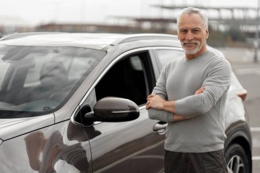 Caucasian handsome gray-bearded elderly man driver, smiles at camera, rejoices at his newly bought luxury car, standing with arms crossed in the dealership parking. Driving People Lifestyle Success