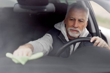 Caucasian senior man 60-70 years old, sitting on the driver's seat and keeping his clean, applying wax in the dashboard and wiping the interior of his car. Automobile service and car maintenance
