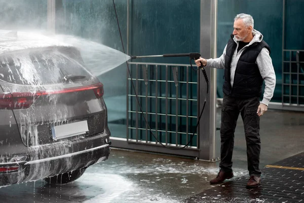 Full-length portrait of elderly Caucasian white-bearded man, washing off the detergent foam soap suds from his car with water from pressurized hose, at a self-service car wash station. Car maintenance