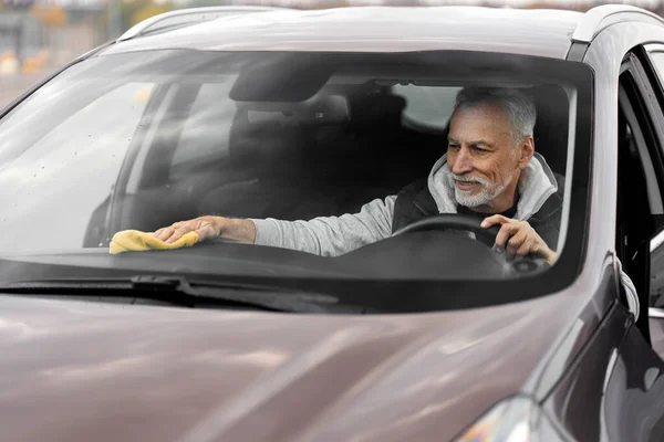 View through windshield to gray-haired bearded Caucasian 60-70 years old man, sitting on driver\'s seat, wiping the car interior with a microfiber. Car wash, maintenance, detailing and valeting concept