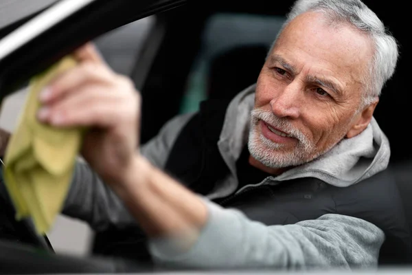 Close-up portrait of handsome positive white-bearded Caucasian 60-70 years old man, sitting on the driver\'s seat and wiping the car\'s side mirror. Car wash and auto service concept