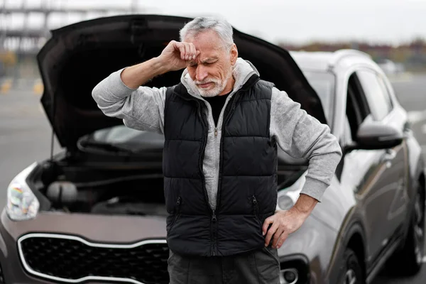 Perplexed anxious senior man driver, feeling emotional stress, holding hand on head, standing near his broken down auto with open hood, on road. Roadside assistance and car insurance. Engine failure