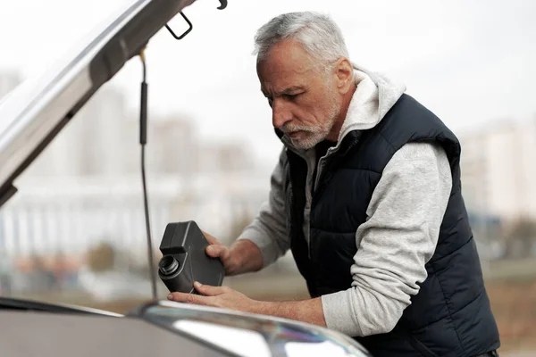 60-70 years old elderly man driver standing by his car with open hood, checking the oil level, pouring and refueling lubricant oil of high quality into the engine motor. Auto service. Car maintenance