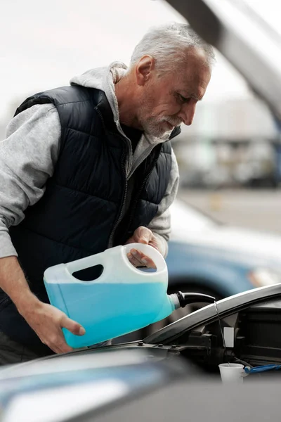 Close-up elderly man, 60-70 years driver, standing by his land vehicle with open hood, filling the reservoir with blue fluid windscreen washer. The concept of car maintenance