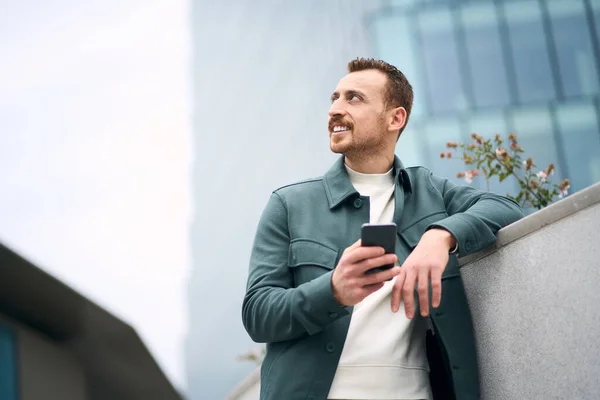 Attractive smiling man using smart phone chatting looking at camera standing on the street