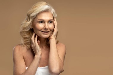 Kendine güvenen güzel beyaz, olgun, doğal makyajlı bir kadının portresi. Yüzüne nazikçe dokunuyor, bej renkli arka planda tek başına poz verirken yan bakıyor.