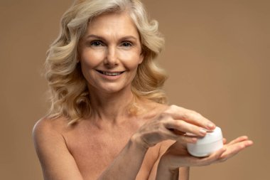 50 'li yaşlarda çekici, olgun, elinde nemlendirici ile kavanoz tutan kameraya gülümseyen bir kadının bej arka planında izole edilmiş portresi. Güzellik tedavisi. Yaşlanma karşıtı, yumuşatıcı, gençleştirici tedavi