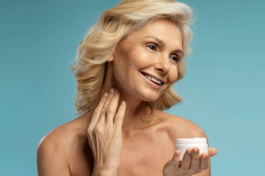 Gülümseyen beyaz, orta yaşlı, güzel, yetişkin bir kadın. Yaşlanma karşıtı kremler sürüyor. Yüzünde nemlendirici ile pürüzsüz kırışıklıklar, mavi arka planda izole edilmiş. Cilt bakımı kavramı