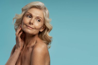 Güzellik tedaviden sonra mükemmel cildine dokunan, nemlendirici süren, reklam metnin için bir kenara bakan, mavi arkaplanda izole edilmiş, muhteşem beyaz yaşlı kadın.