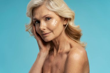 Güzel, kendine güvenen, beyaz, olgun, sağlıklı cilde sahip, nemlendirici sürmüş, mavi arka planda izole edilmiş, reklam alanı olan bir kadın. Deri ve vücut bakımı, olgun güzellik konsepti