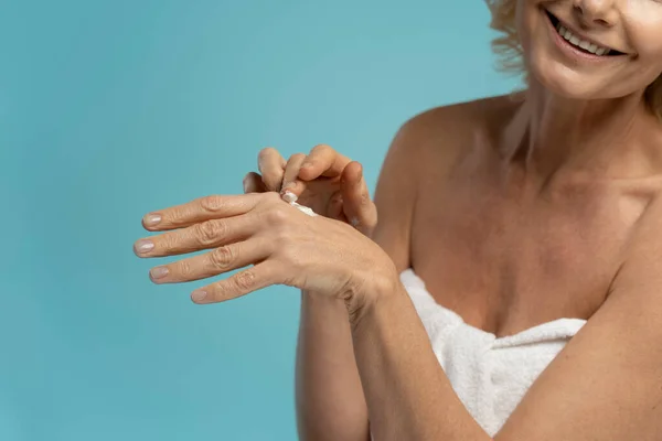 stock image Cropped view of Caucasian smiling mature woman wrapped in towel, applying moisturizing cream on her hands on blue background with copy space. Body and skin care. Morning routines. Spa beauty procedure