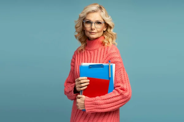 迷人的白种人成熟女人 金发碧眼 戴着眼镜 穿着温暖的粉色毛衣 摆设着便笺和课本 凝视着蓝色背景的相机 — 图库照片
