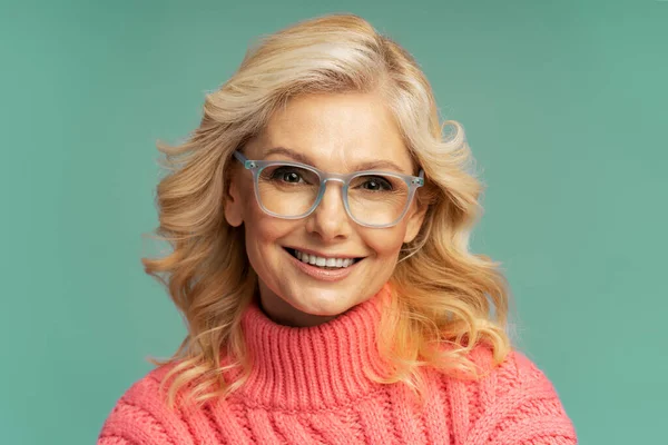 Isolated close-up portrait on colored background of a confident charming Caucasian mature woman with beautiful wavy blond hair, wearing eyeglasses, smiling a beautiful toothy smile, looking at camera
