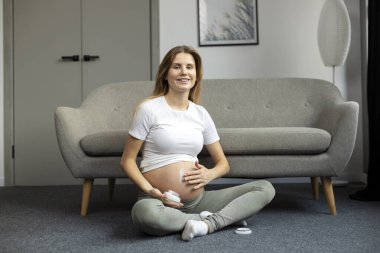 Beautiful smiling pregnant woman applying stretch mark cream to belly while sitting at home. Pregnancy, skin care people and maternity concept 