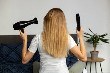 Back view of the blonde woman drying and styling hair with hairdryer while making hairstyle at home. Happy female using hair dryer and enjoying beauty routine 