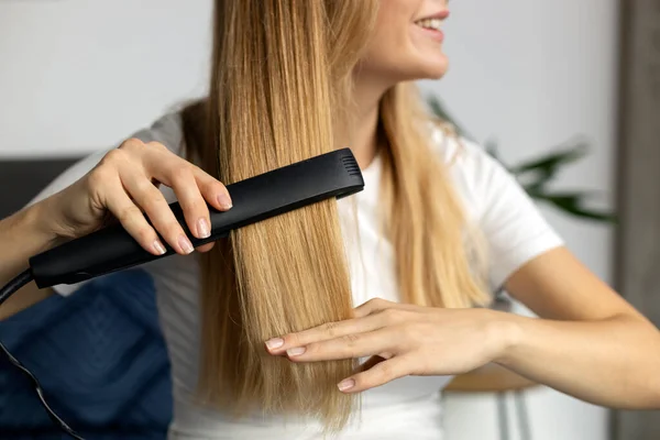 Beautiful smiling woman using hair straightener at home, selective focus on hair. Beauty, hair care concept