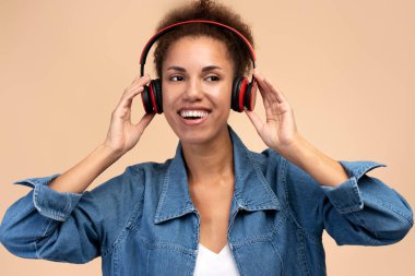 Beautiful multi-ethnic woman with afro hairstyle, dressed in blue denim casual shirt, looking aside while singing song, wearing wireless headphones, isolated on beige background. People. Lifestyle
