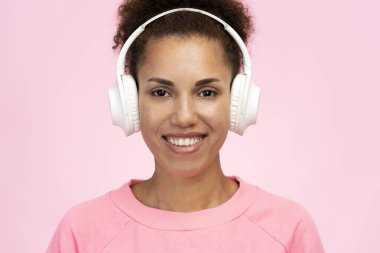 Close up portrait of confident African American delightful woman, wearing wireless headphones and pink sweatshirt, smiling beautiful toothy smile looking at camera, isolated on pink pastel background