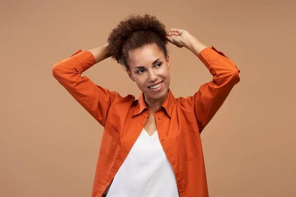 Beautiful African American woman in orange shirt and white t-shirt, smiling looking aside, fixing her stylish afro hair, expressing positive emotion, isolated over beige background. People. Lifestyle.