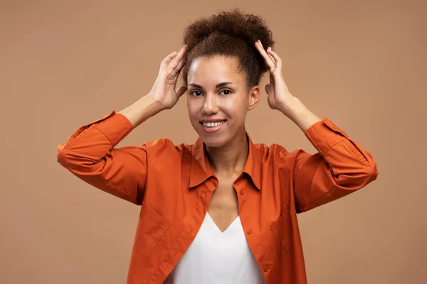 Stunning woman with afro hair, wearing orange casual shirt and white t-shirt, smiling a cheerful smile looking at camera, expressing positive emotions, isolated beige background. People. Lifestyle.