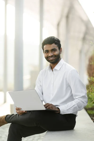 Smiling handsome Indian programmer using laptop computer working online at workplace. Portrait of happy businessman watching training courses, planning startup. Successful business concept