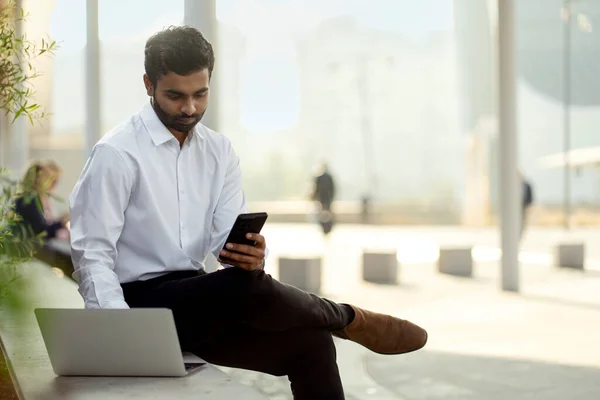 Portrait of pensive handsome Indian businessman using laptop computer holding smartphone working outdoors. Confident asian freelancer check email, communication online, planning project at workplace