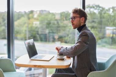 Multitasking young male entrepreneur, businessman in formal suit, looking away, checking time on wrist watch while waiting for people at job interview while sitting at laptop in modern light office