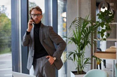 Confident young businessman, wearing elegant formal suit and eyeglasses, talking on mobile phone, negotiating with investors or business partners while standing by the window in modern office interior