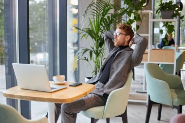 Young man, freelance entrepreneur, office worker wearing stylish business suit and eyeglasses, relaxing while taking a coffee break, sitting at a desk with laptop in a cozy coffee shop interior.